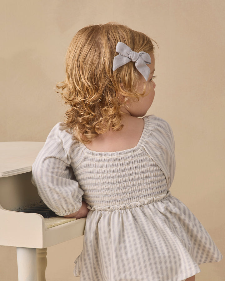 A toddler with curly blonde hair wearing a light gray bow headband and a smocked long-sleeve dress with thin vertical stripes, leaning on a white piano against a beige background.