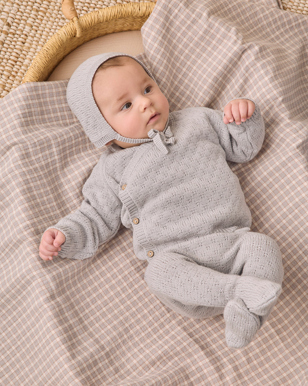 A baby wearing a light gray knit bonnet and matching footed one-piece outfit lies on a beige and white checkered blanket next to a woven basket.