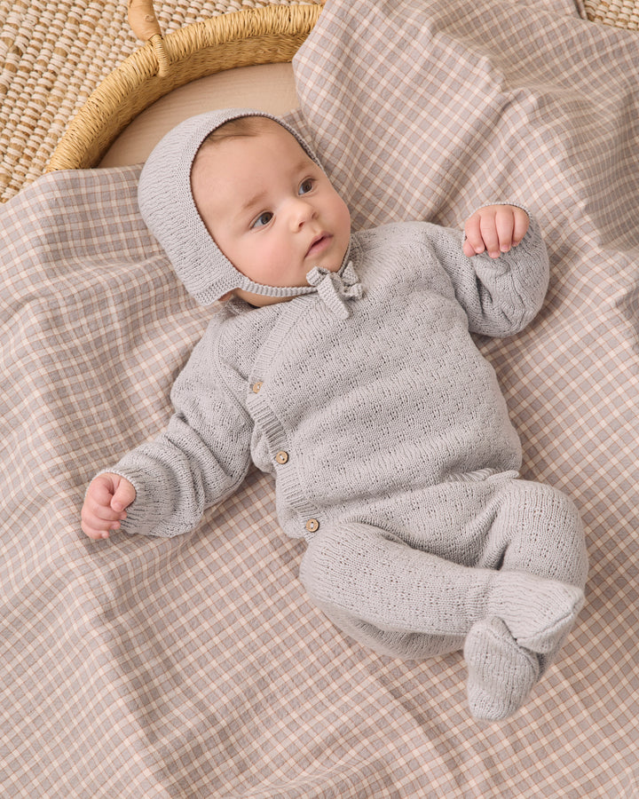 A baby wearing a light gray knit bonnet and matching footed one-piece outfit lies on a beige and white checkered blanket next to a woven basket.