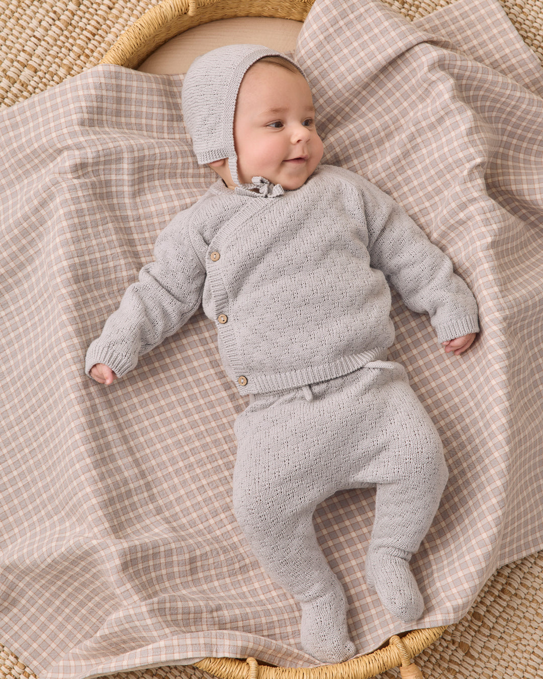 A baby wearing a light gray knit cardigan, matching footed pants, and a bonnet, lying on a beige and white checkered blanket in a woven basket.