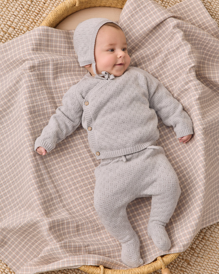 A baby wearing a light gray knit cardigan, matching footed pants, and a bonnet, lying on a beige and white checkered blanket in a woven basket.