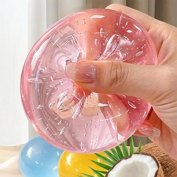 A hand holding a translucent pink squishy gel disc with a textured, bubbly surface, shown against a softly blurred indoor background with colorful props.