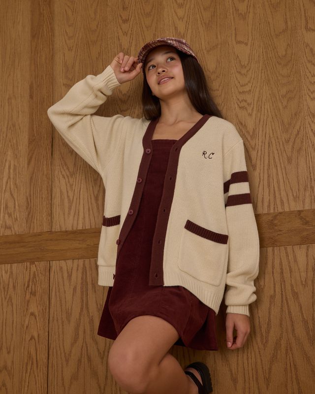 Woman wearing a beige cardigan over a maroon dress with a matching hat against a wooden paneled wall.