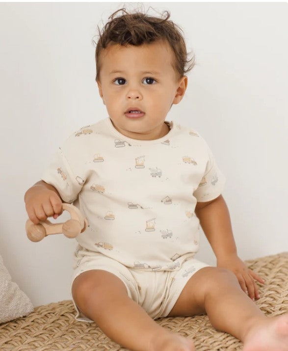 Baby sitting on a woven surface holding a wooden toy, wearing a light-colored outfit with patterns.