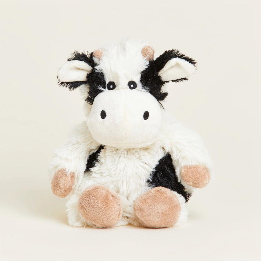 A plush toy cow with fluffy white and black fur, pinkish-tan hooves, and small black eyes, sitting against a plain light beige background.