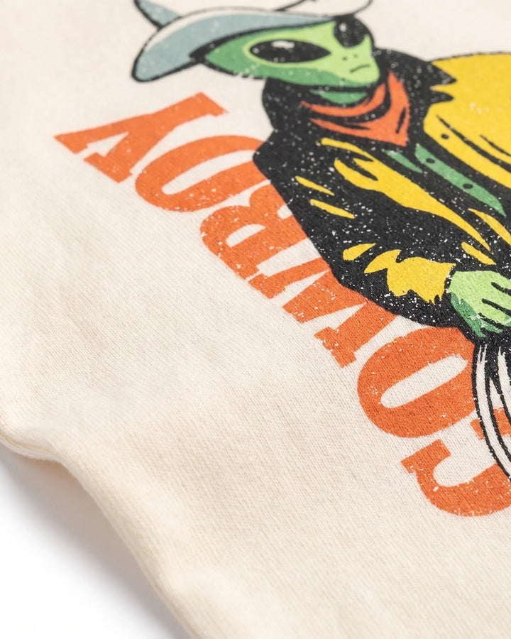 Close-up of a cream-colored t-shirt featuring a retro graphic of a green alien wearing a cowboy hat and vest, with the word 'COWBOY' printed in large orange letters.
