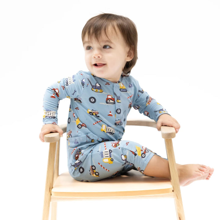 A toddler sitting on a small wooden chair wearing a light blue long-sleeve pajama set with a colorful construction vehicle print, photographed against a clean white background.