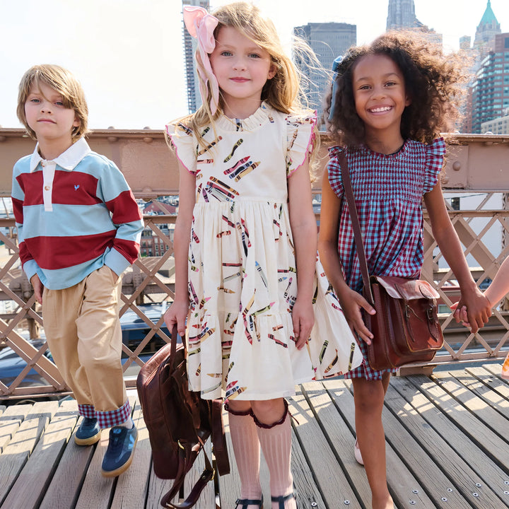 Three children modeling fashion on a wooden bridge with a city skyline in the background. The central girl wears an off-white dress with a colorful pencil print and a pink bow, holding a brown leather bag.
