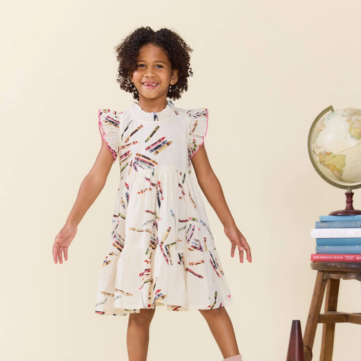 A young girl with curly dark hair stands smiling in a cream-colored dress decorated with a colorful pencil print. She is posed in front of a plain beige background with a stack of books and a globe on a small wooden stand to the right.