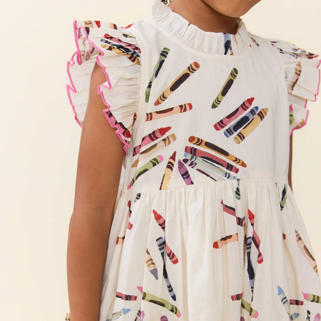 A close-up of a child wearing a white ruffled dress with a colorful crayon print and pink-trimmed flutter sleeves, shown against a plain light beige background.
