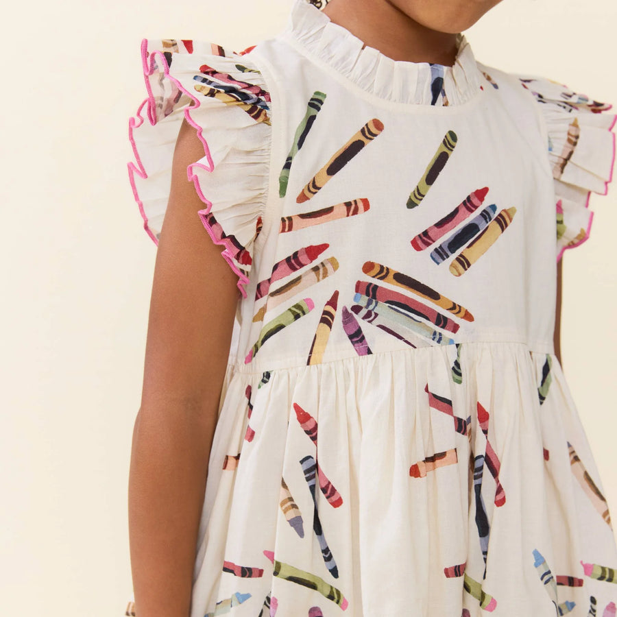 A close-up of a child wearing a white ruffled dress with a colorful crayon print and pink-trimmed flutter sleeves, shown against a plain light beige background.