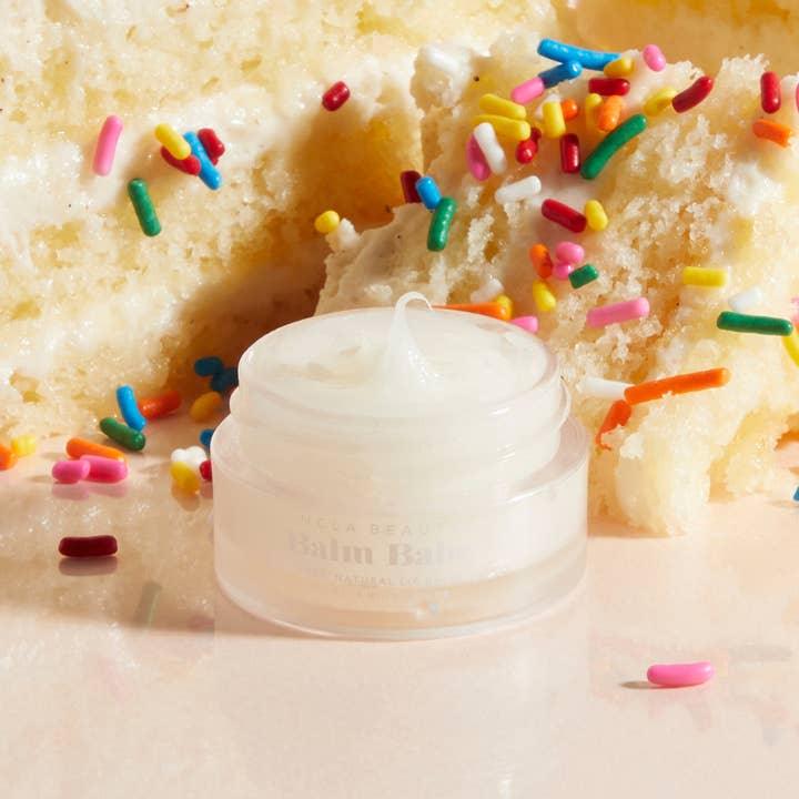 PIcture of the lip balm near a piece of vanilla cake with sprinkles to celebrate the scent of the lip balm
