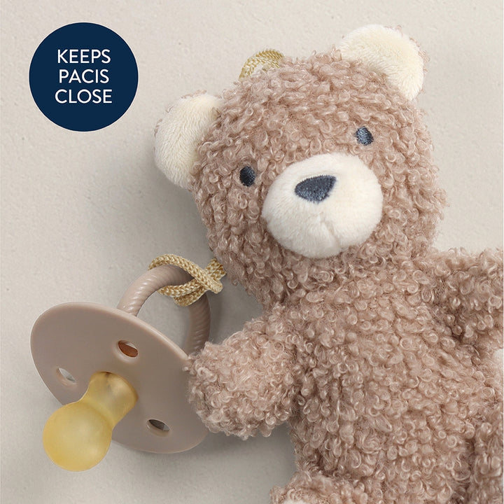 Teddy bear holding a pacifier with a text label 'Keeps Pacis Close' on a light background