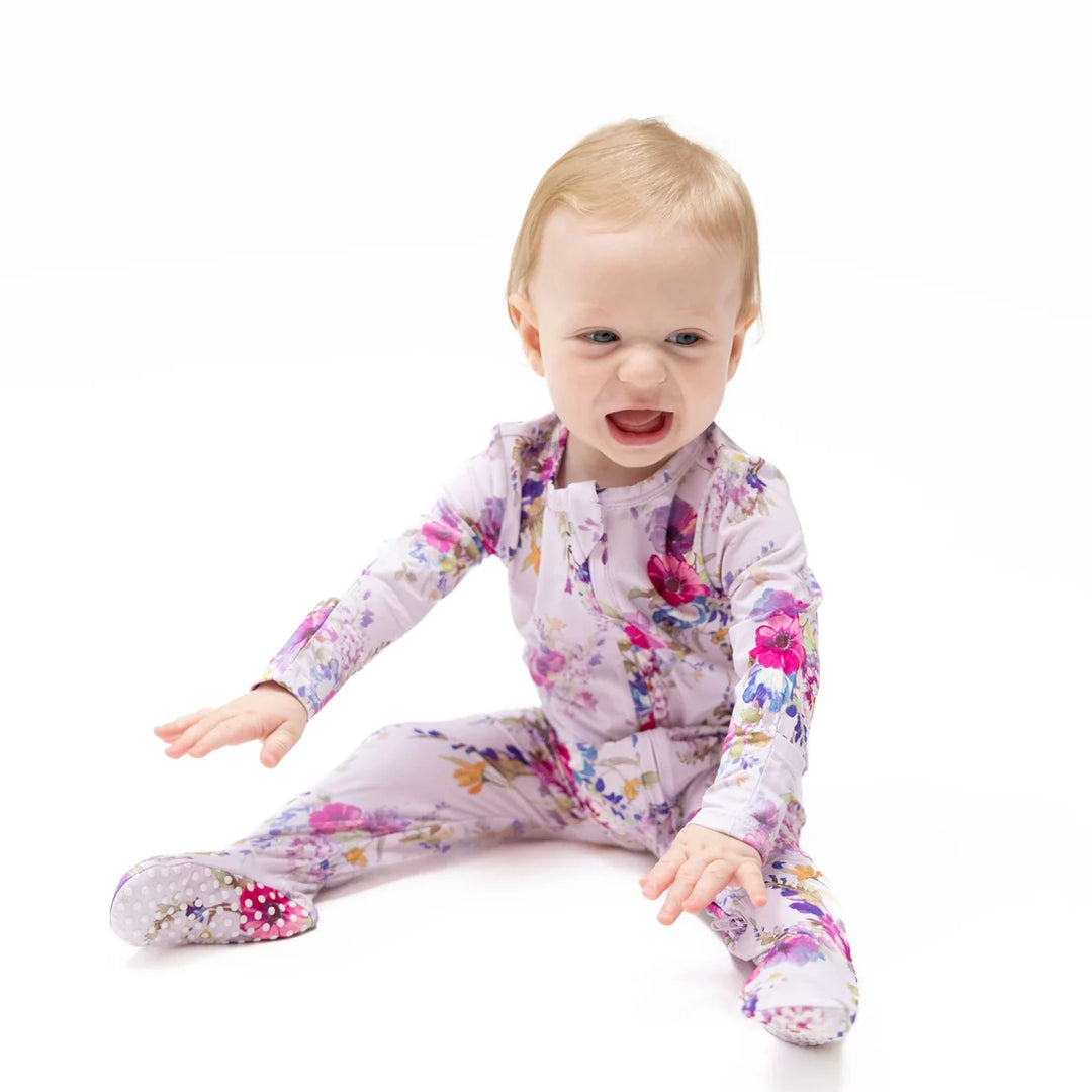 A baby sitting on a white studio background wearing a light lavender footed sleeper with a colorful pink, purple, and blue floral print.