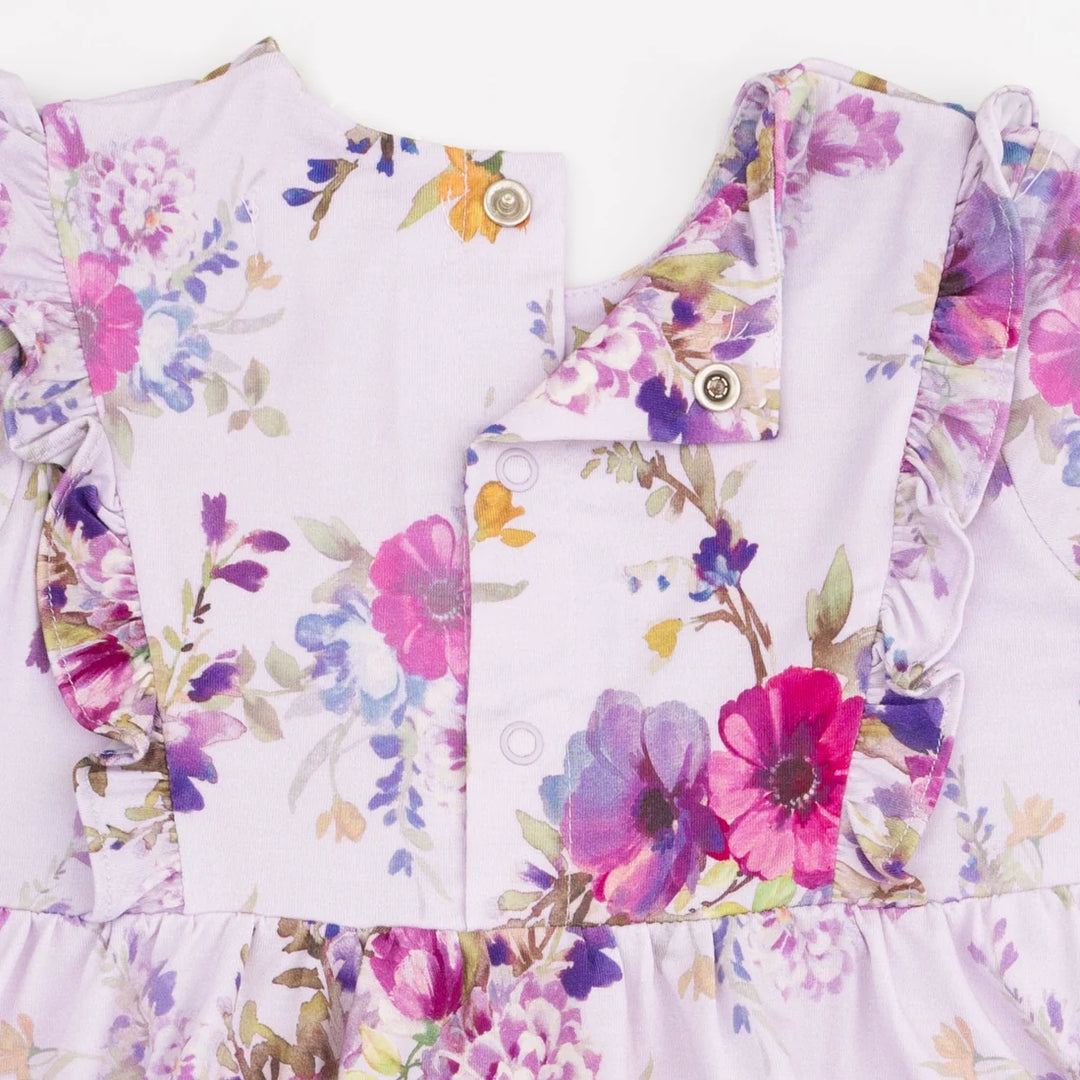 Close-up of a children's dress or top with a light lavender base and a colorful floral print in pink, purple, blue, and green. The garment features ruffled trim, a buttoned front panel, and decorative buttons.