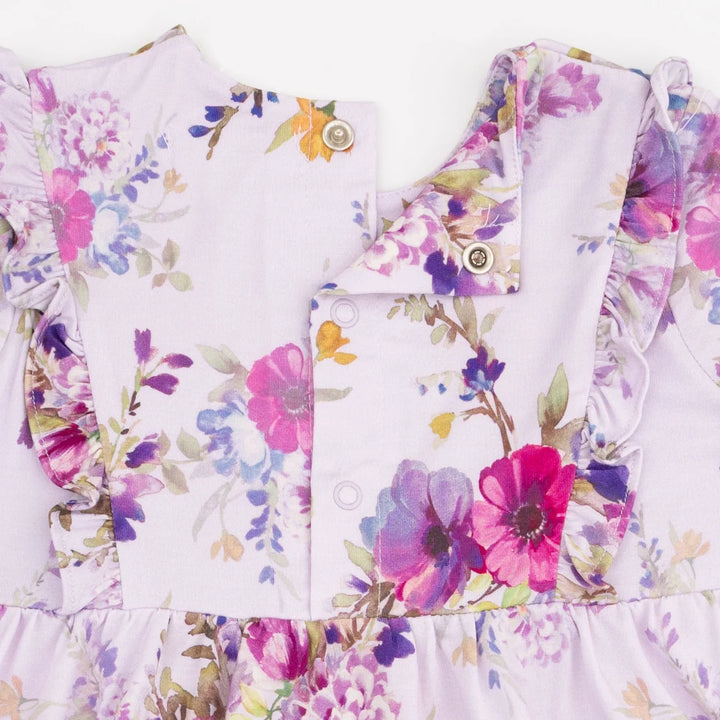 Close-up of a children's dress or top with a light lavender base and a colorful floral print in pink, purple, blue, and green. The garment features ruffled trim, a buttoned front panel, and decorative buttons.