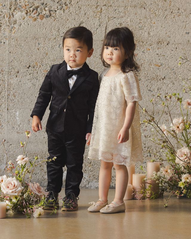 Two children, a boy in a suit and a girl in a dress, standing in front of a floral arrangement.