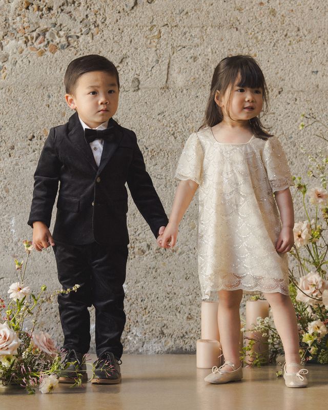 Two children, a boy in a suit and a girl in a dress, holding hands against a textured wall with floral decorations.