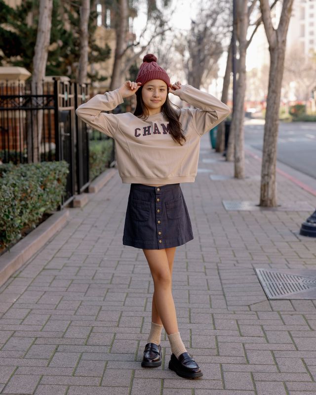 Woman wearing a beige sweatshirt with 'CHAMP' branding, dark denim skirt, and black loafers on a sidewalk.