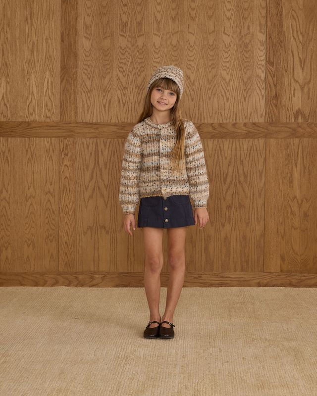 Young girl wearing a patterned sweater and denim skirt against a wooden paneled wall.
