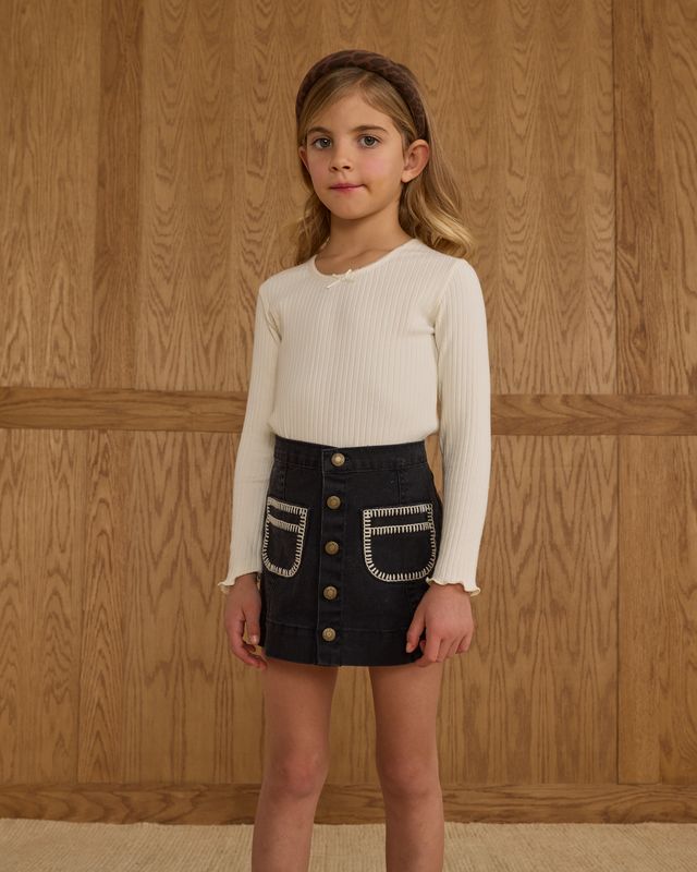 Young girl wearing a white blouse and black skirt with button details against a wooden paneled wall.