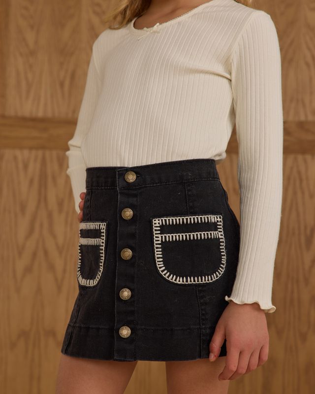 Person wearing a white ribbed sweater and black denim skirt with gold buttons and embroidery on a wooden background