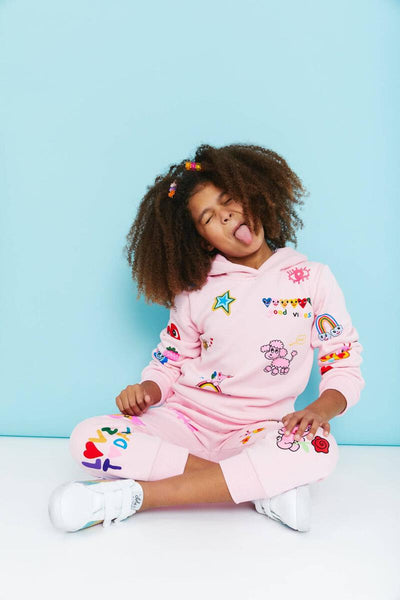 Little girl modeling the hoodie 2 piece set. pink joggers and pink hoodie with all over doodle print.  little girl has her tongue sticking out and is sitting down. 