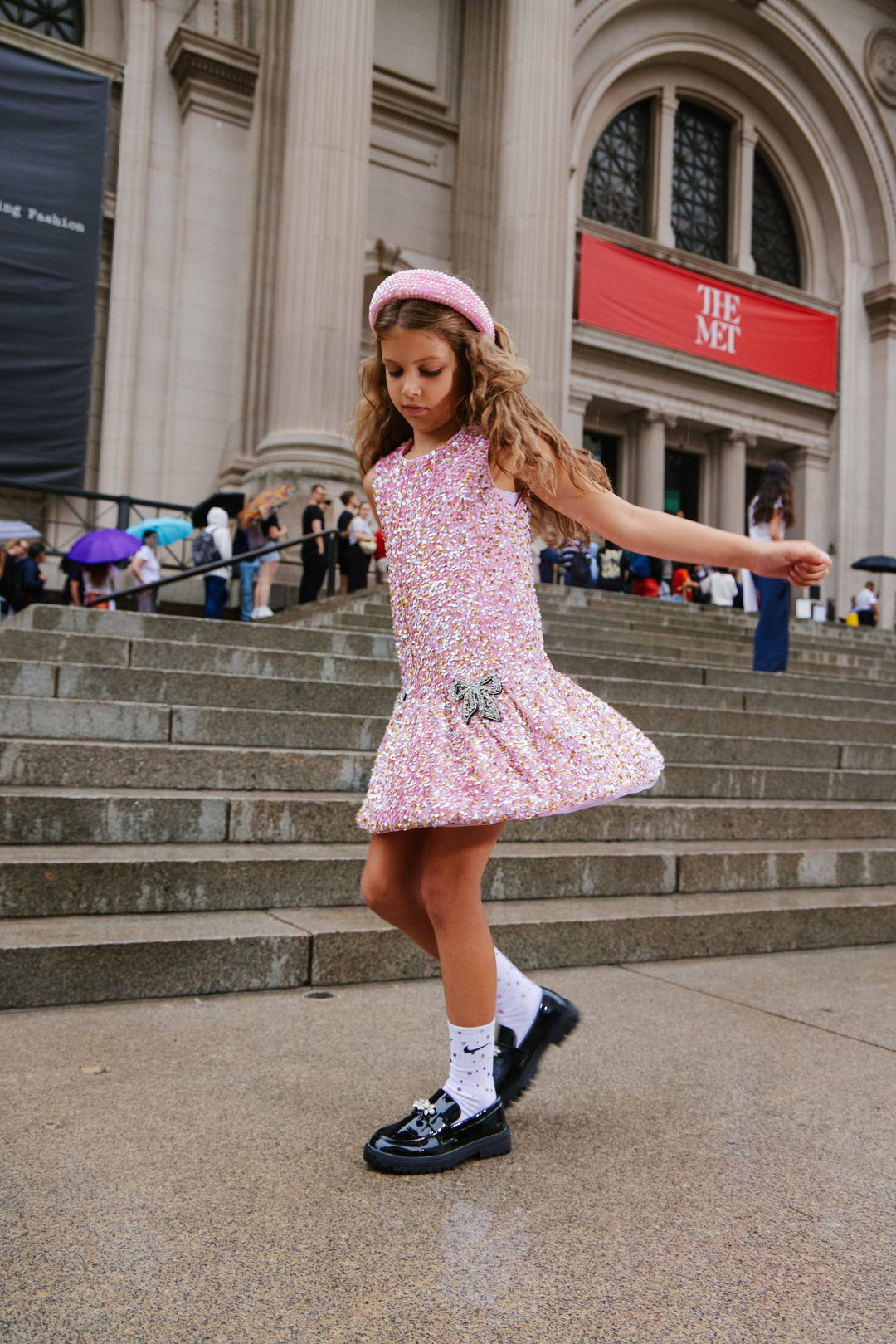 Young girl in a pink dress standing on steps in front of a large building with a red sign.