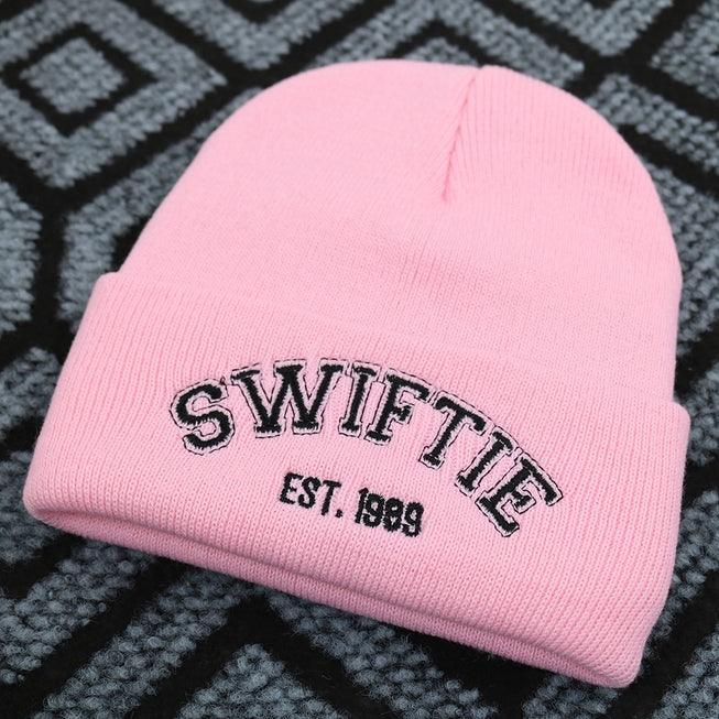 light pink beanie with the word 'Swiftie' written across it in black thread. 