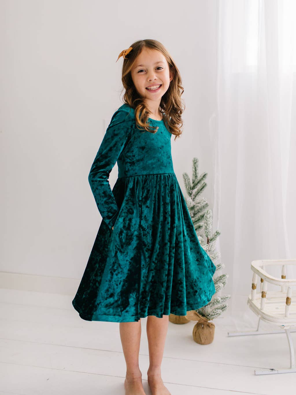 Young girl wearing a deep green dress standing in a bright room with a small Christmas tree.