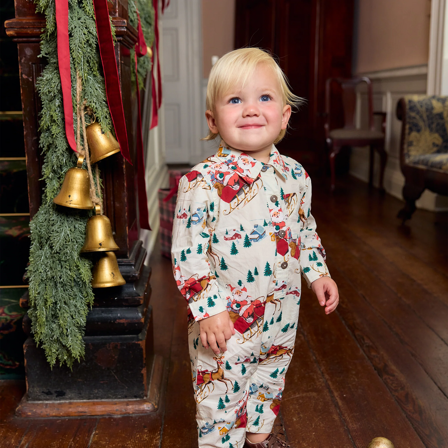 A young child wearing a festive Christmas-themed long-sleeve pajama set with a printed pattern of Santa Claus, reindeer, snow globes, and Christmas trees, standing in a decorated indoor hallway with wooden floors and holiday garlands.