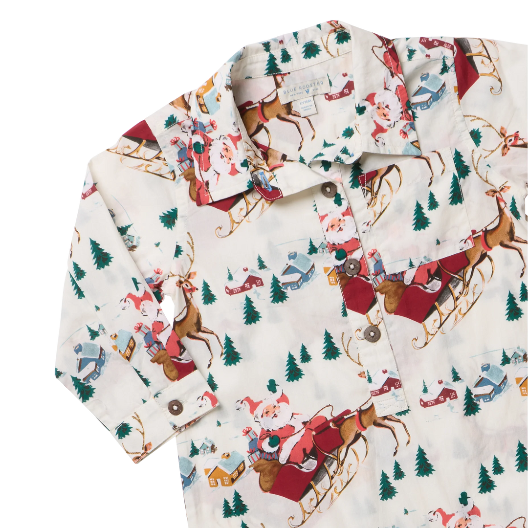 A long-sleeve button-up shirt with a festive Christmas print featuring Santa Claus in a sleigh, reindeer, pine trees, and small houses on an off-white background.