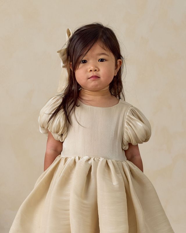 Young girl wearing a beige dress with puffed sleeves against a plain background