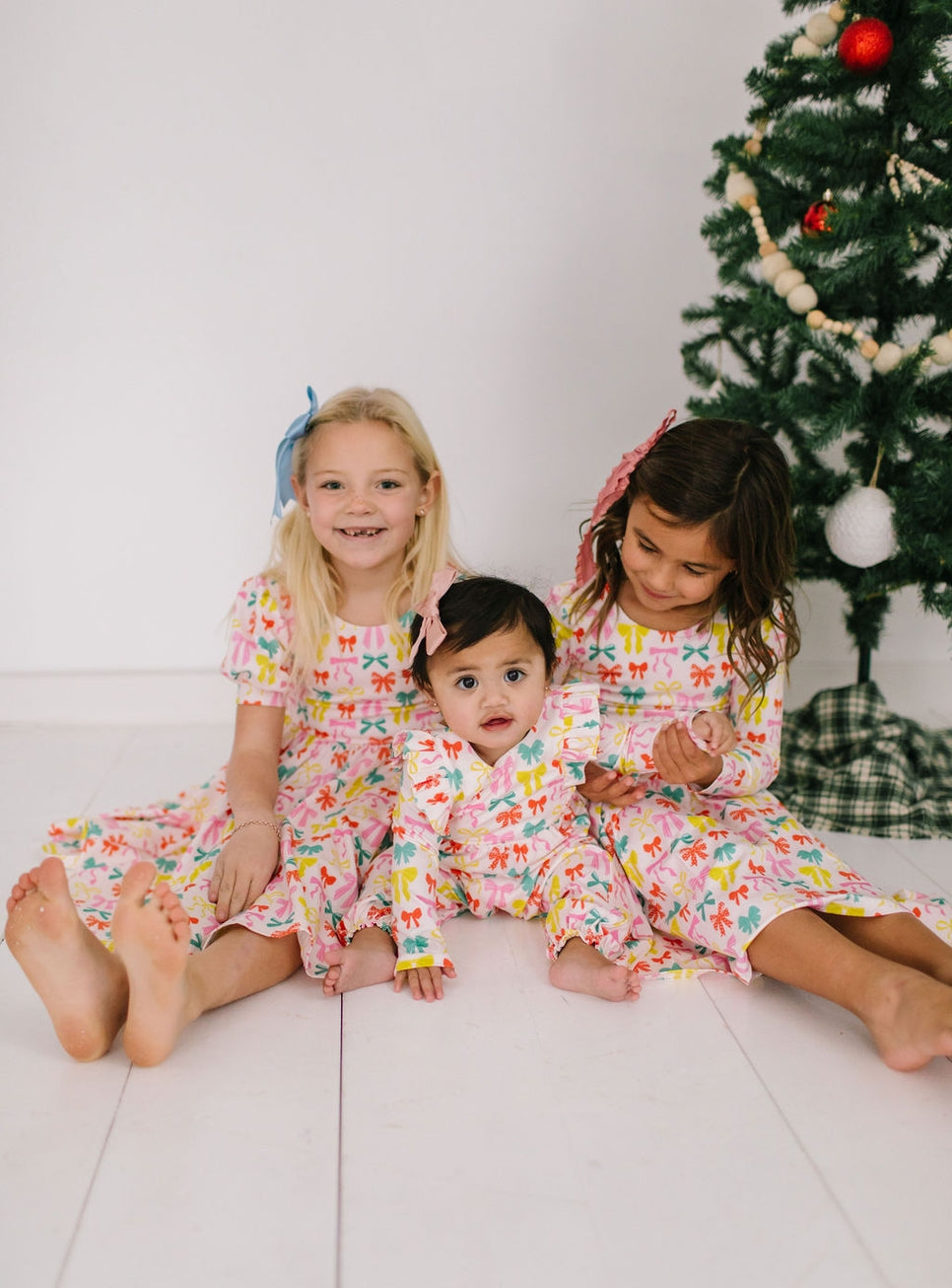 Three children in matching printed dresses with colorful bows on them sitting on a white floor with a decorated Christmas tree in the background.