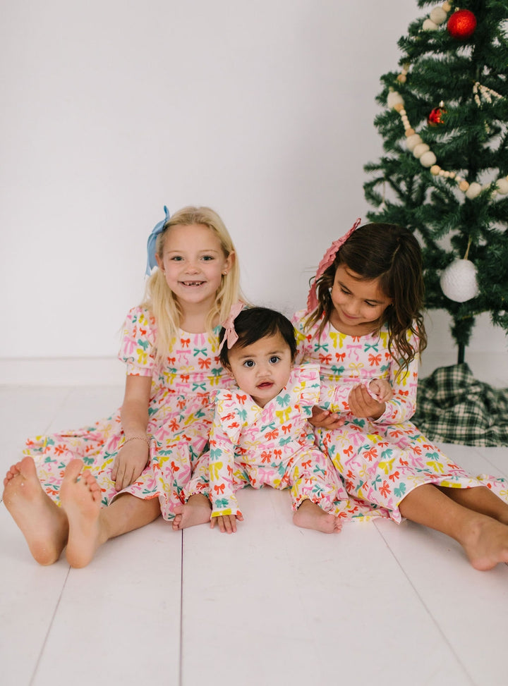 Three children in matching printed dresses with colorful bows on them sitting on a white floor with a decorated Christmas tree in the background.