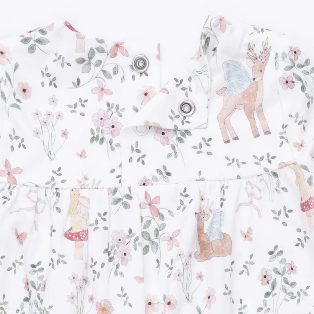 A close-up of a white baby romper featuring a whimsical fairy and deer print with pink flowers, green leaves, and small mushrooms. The dress has a ruffled waist and snap-button closures at the neckline.