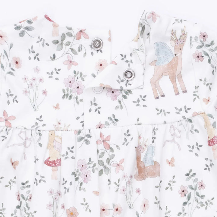 A close-up of a white baby romper featuring a whimsical fairy and deer print with pink flowers, green leaves, and small mushrooms. The dress has a ruffled waist and snap-button closures at the neckline.