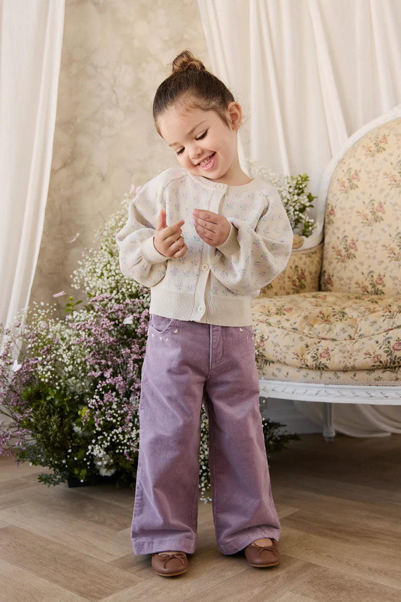 Young girl in a light sweater and purple pants standing in a room with flowers and a sofa.
