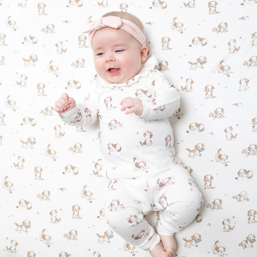 A baby lying on a patterned dog-print blanket, wearing a white long-sleeve footed sleeper with a ruffled collar and a matching pink bow headband.