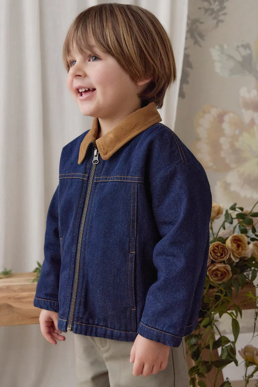 A young boy wearing a dark blue denim jacket with a tan corduroy collar, standing in a softly lit indoor setting with curtains and flowers in the background.