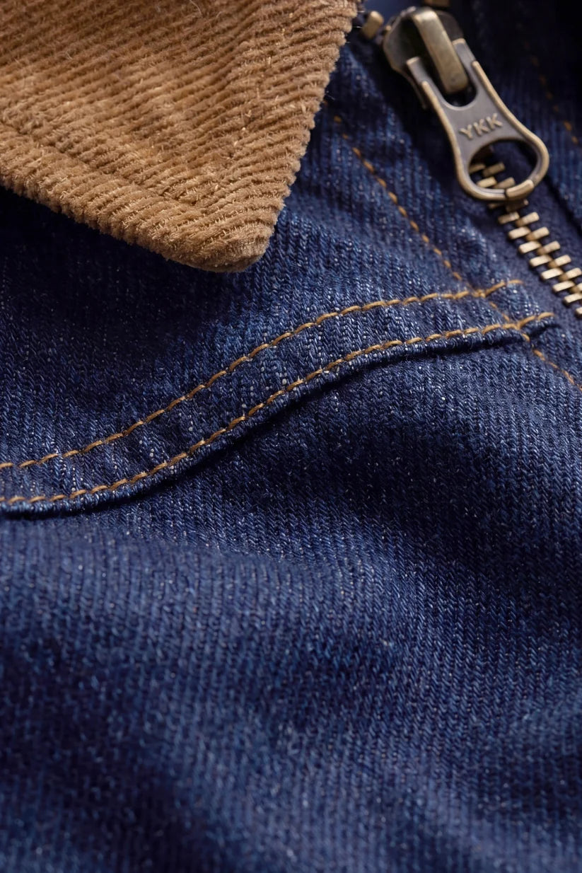 Close-up detail of a dark blue denim jacket featuring a brown corduroy collar, gold contrast stitching, and a silver metal zipper.