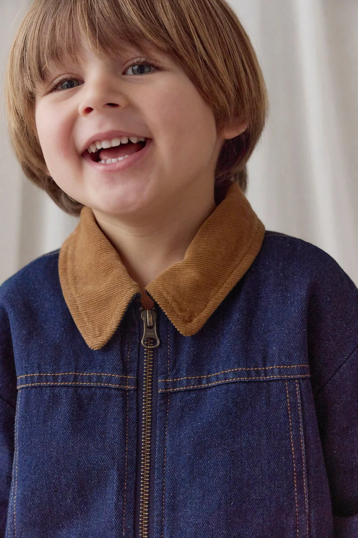 A close-up of a young boy smiling, wearing a blue denim-style jacket with a brown corduroy collar.