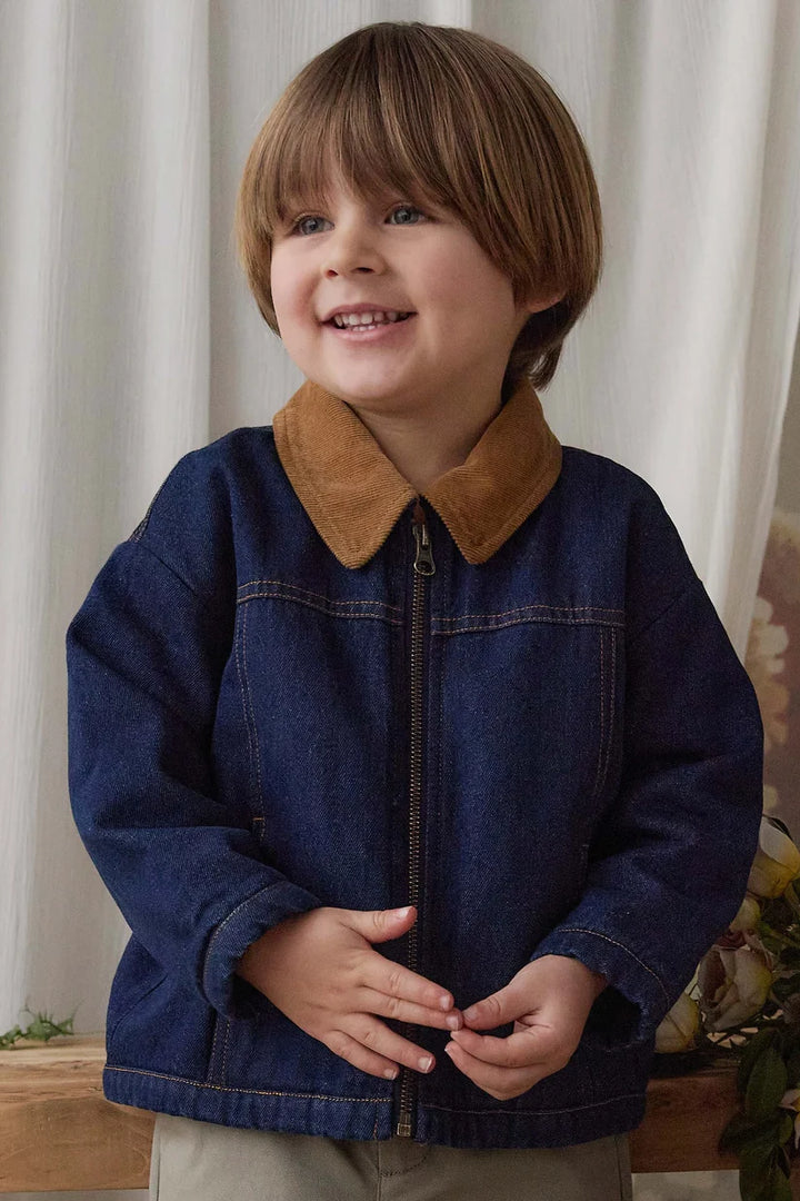A young boy with brown hair smiling while wearing a dark blue denim jacket with a brown suede collar, standing indoors against a light curtain background.