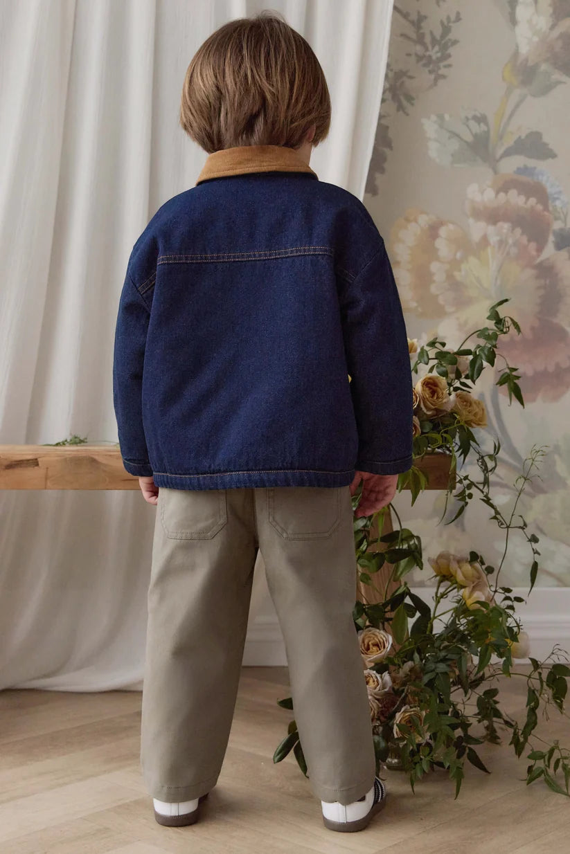 A child viewed from behind wearing a navy blue denim jacket with a tan collar, paired with beige trousers, standing in a room with floral wallpaper, a wooden bench, and curtains.