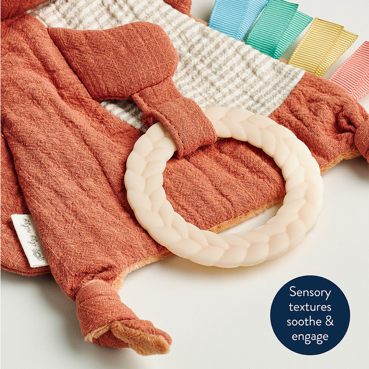 Textured orange baby garment with a white ring on a patterned surface, featuring a label and text about sensory textures.