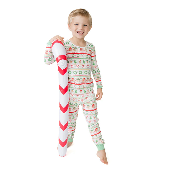 A young child wearing a festive holiday-themed long-sleeve pajama set stands barefoot against a white background, holding a large white candy cane prop. The pajamas feature a repeating Christmas pattern with red, green, and white colors.