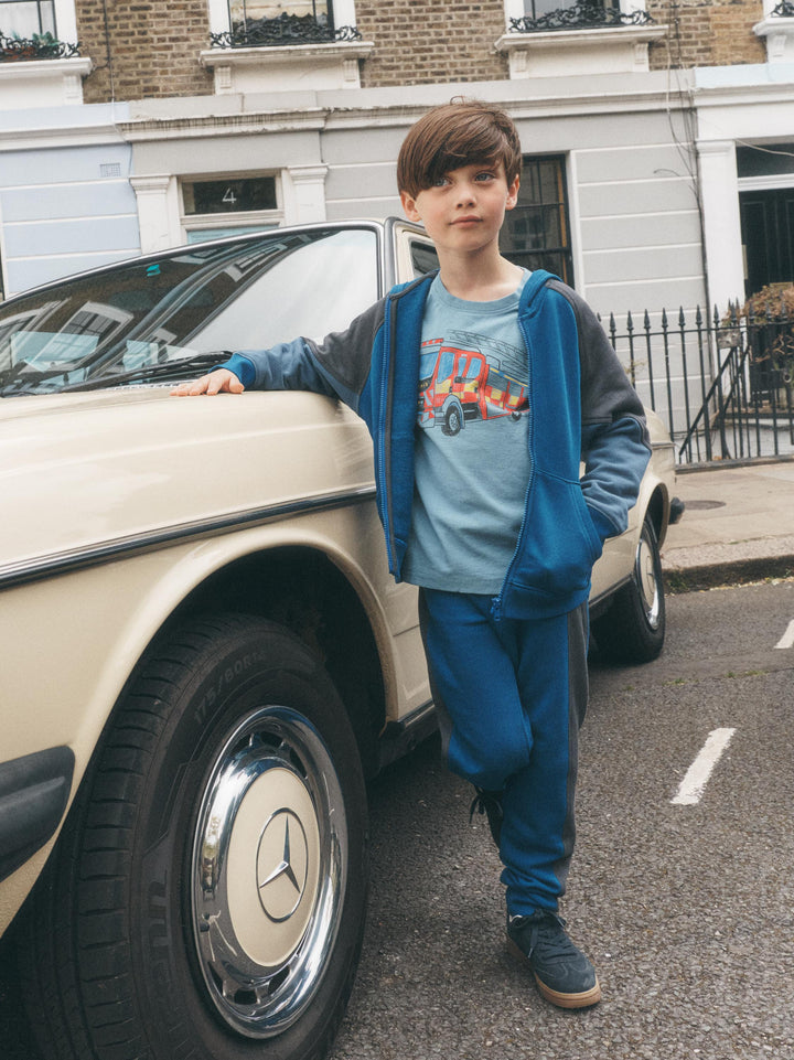 A young boy stands on a street next to a cream-colored classic Mercedes-Benz car, wearing a blue graphic t-shirt, a blue zip-up hoodie, and blue sweatpants.