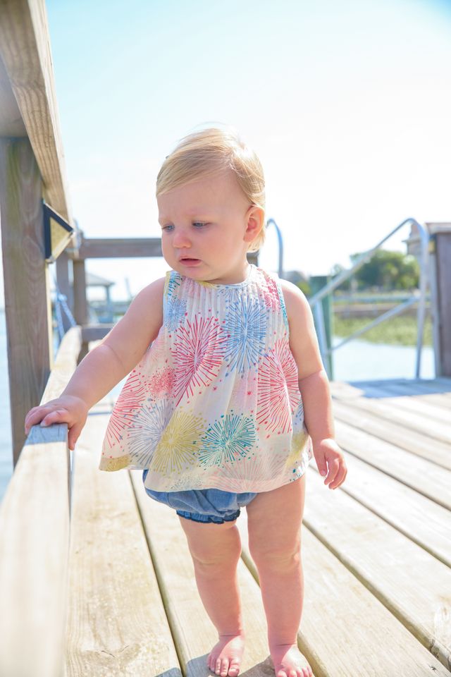 Baby standing on a wooden deck wearing a colorful sleeveless top and denim shorts.