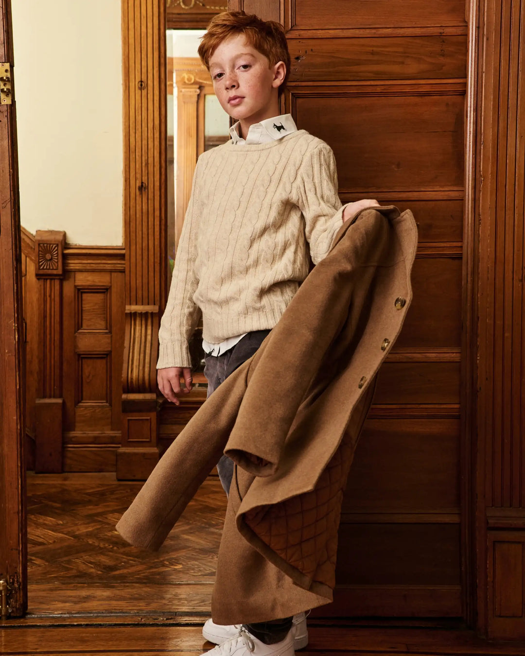 A young boy stands in a warm, wood-paneled room holding a long brown coat. He is wearing a cream cable-knit sweater over a white collared shirt, gray jeans, and white sneakers.