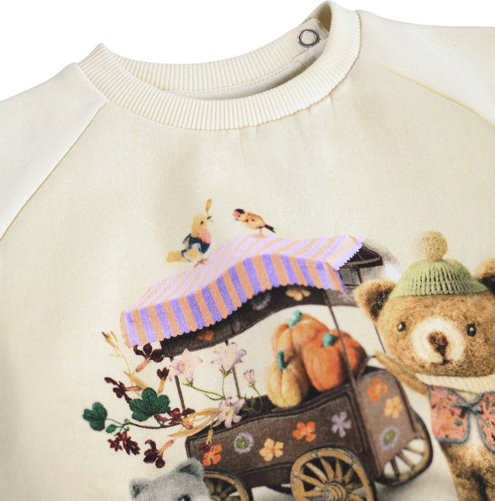 Close-up of a cream-colored raglan sweatshirt featuring a colorful graphic print of a teddy bear, a pumpkin cart, and autumn flowers.